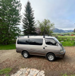 1994 Toyota Hiace SuperCustom van parked outdoors with a scenic background, featuring roof rack and new tires.